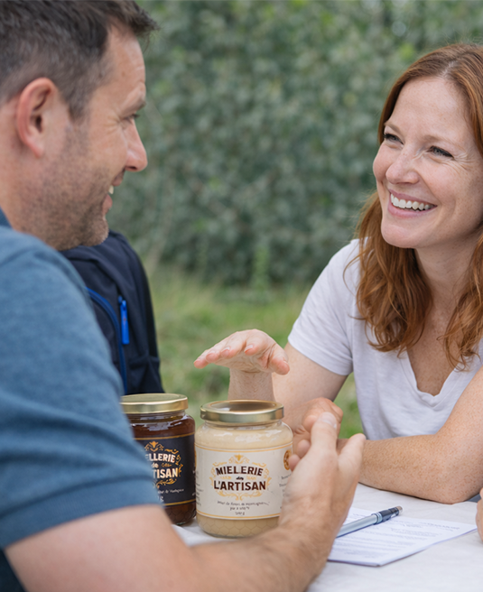 Couple souriant avec pots de miel artisanal "Mielerie l'Artisan" sur table. Signature de contrat possible.